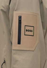 Manche de veste beige avec une poche rectangulaire beige dotée d'une fermeture éclair noire verticale et du logo "BOSS" à l'intérieur d'un cadre carré noir.