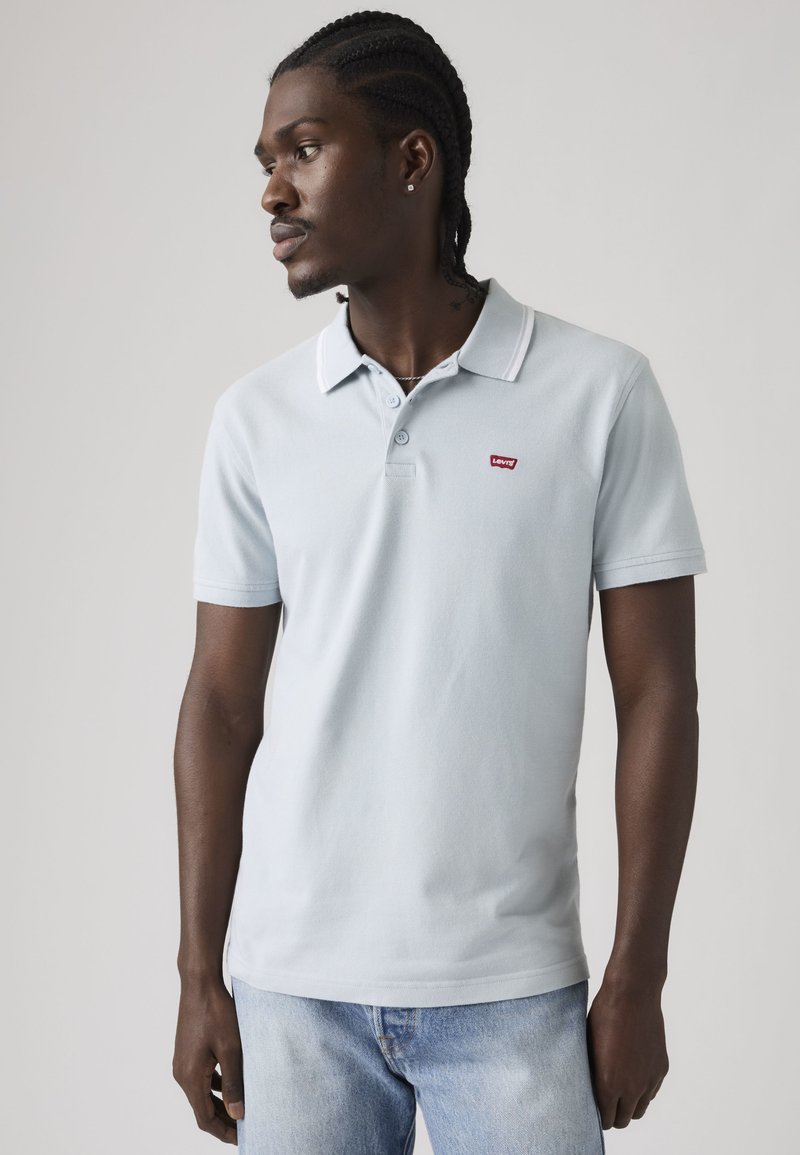 Hombre con el pelo trenzado que lleva una camiseta polo azul claro de Levi's y vaqueros desgastados, mirando hacia su izquierda sobre un fondo liso.