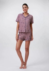 Νεαρή γυναίκα που στέκεται ξυπόλητη, φορώντας σετ πιτζάμας με κοντό μανίκι και σορτς, με ροζ σχέδια, χαμογελώντας προς την κάμερα.