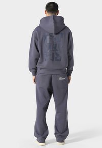 Conjunto de sudadera con capucha y pantalones de chándal gris oscuro, con un gran gráfico en la parte posterior, puños elásticos y un ajuste relajado. Tejido suave y texturizado.