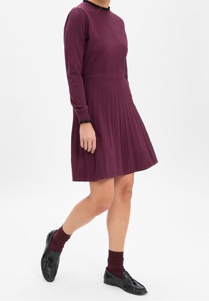 Vrouw draagt een bordeauxrode gebreide jurk met lange mouwen en geplooide rok, bijpassende sokken en zwarte loafers, staand tegen een witte achtergrond.