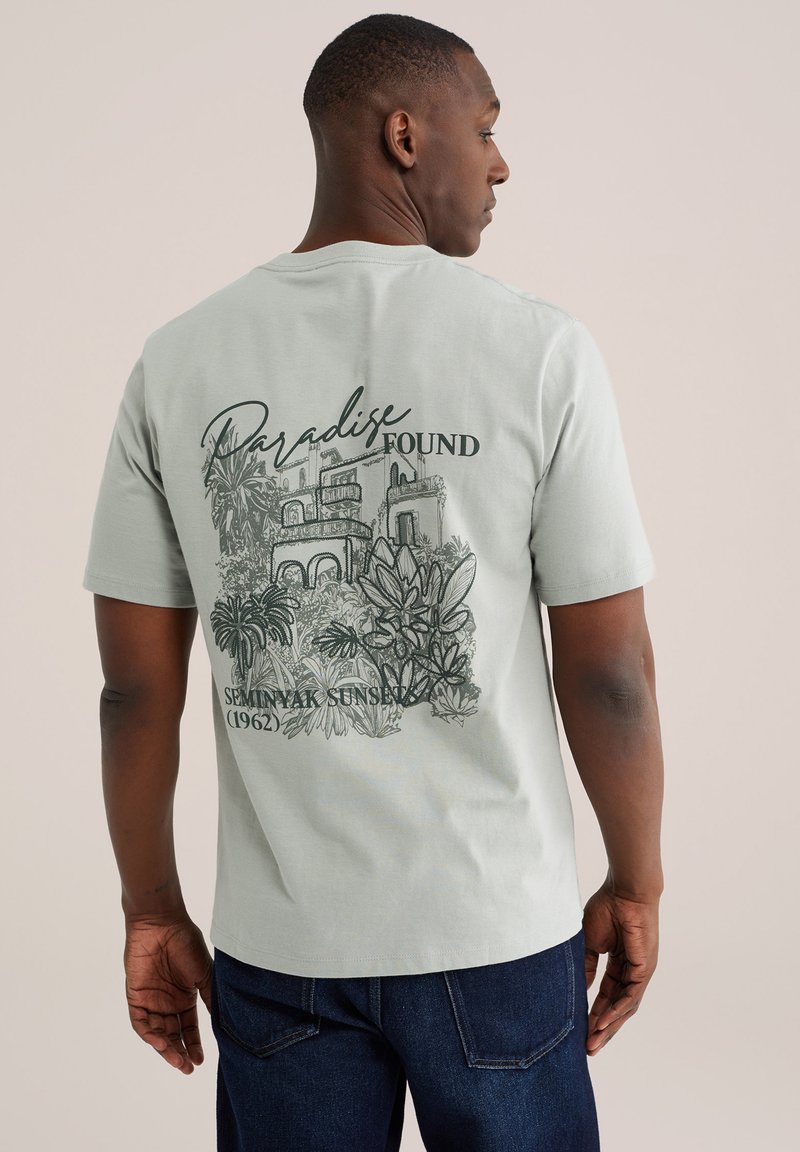 Mann trägt ein hellgraues T-Shirt mit einem Rückendruck von tropischen Pflanzen, Gebäuden und dem Schriftzug "Paradise Found Seminyak Sunset (1962)".