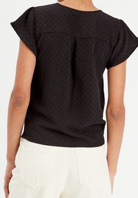 Haut noir à manches courtes en tissu texturé, col rond et détail plissé au dos, associé à un pantalon crème.