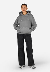 Šedá mikina s kapucňou a predným vreckom, doplnená o logo, v kombinácii s čiernymi džínsami s vysokým pásom. Model nosí biele a šedé tenisky. Jednoduchý dizajn.