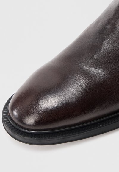 Close-up da pontinha de um sapato de couro polido castanho-escuro com sola de borracha preta texturizada sobre fundo branco.