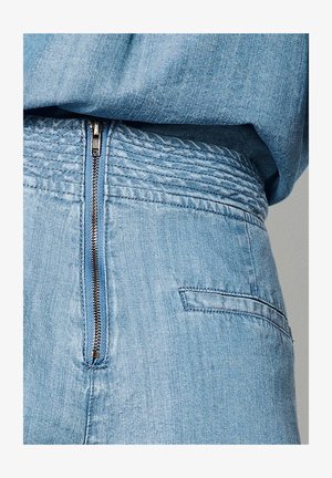 Pantalones de mezclilla azul claro con cierre vertical de cremallera metálica en la parte trasera y un bolsillo horizontal con abertura en el lado derecho.