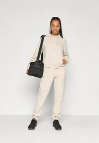 Beżowa bluza z kapturem ze sznurkiem i kieszenią kangurka, zestawiona z dopasowanymi, zwężanymi spodniami dresowymi. Czarna torebka na ramię z tkaniny i czarne trampki.