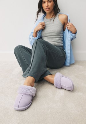 Woman sitting on carpeted floor wearing gray tank top, blue shirt, dark gray pants, and light purple fur-lined slippers.