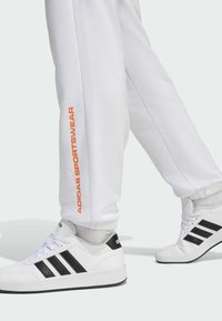 Biele tepláky s nápisom "ADIDAS SPORTSWEAR" v oranžovej farbe po strane, skombinované s bielymi teniskami s čiernymi akcentmi a pruhmi.