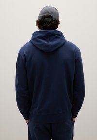 Felpa blu marina con cappuccio regolabile, polsini e orlo a coste; indossata con pantaloni blu marina abbinati, con un cappellino grigio.