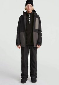 Kurtka narciarska w kolorze czarnym i szarym, z zamkiem błyskawicznym z przodu, kieszeniami naszytymi oraz kapturem. Połączona z czarnymi spodniami i czarnymi butami narciarskimi.