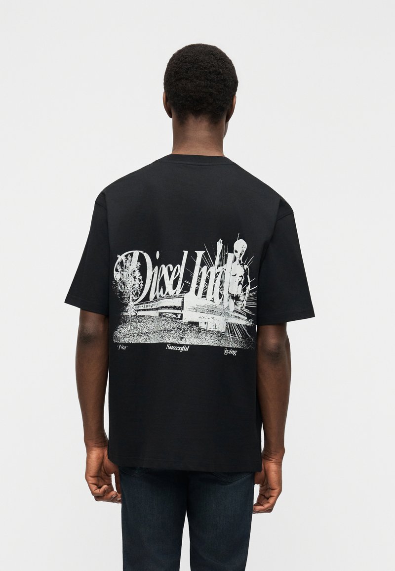 Άνδρας που φοράει μαύρο μπλουζάκι με λευκό γραφικό ενός κτιρίου, δέντρου, σκελετού και κειμένου "Diesel Ind." στην πλάτη, στέκεται μπροστά σε απλό φόντο.