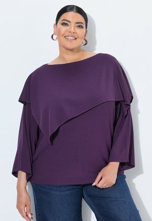 Kvinde med mørkt hår trukket tilbage iført lilla asymmetrisk langærmet top og blå jeans, smilende mod en hvid baggrund.