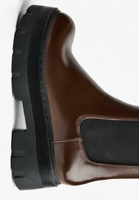 Ghetă maro din piele, cu talpă neagră din cauciuc, având o pană elastică laterală și o textură netedă; vârf rotund și detalii minime de cusături.