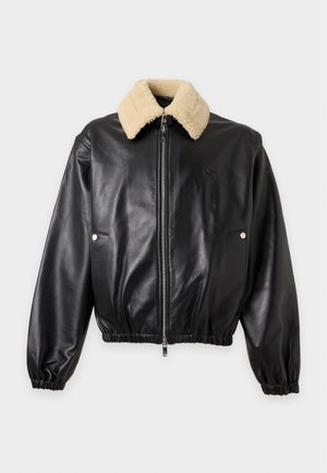 Jeune homme debout portant une veste en cuir noire avec un col en shearling crème, une chemise noire, un jean large gris foncé et des chaussures noires.