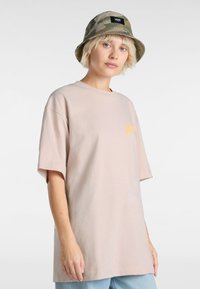 Helles rosa Baumwoll-T-Shirt mit kurzen Ärmeln, das ein kleines gelbes Logo auf der Brust trägt. Kombiniert mit einem camouflagefarbenen Fischerhut.