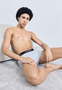 Boxer grigi con una cintura nera "LACOSTE", abbinati a un busto nudo, seduto su un morbido tappeto accanto a una superficie chiara e strutturata.