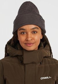 Browns pufīgs mētelis ar augstu apkakli, apvienots ar tumši pelēku ribotu beanie cepuri. Uz mēteļa ir O'Neill zīmols. Mīksta tekstūra, ikdienišķs dizains.
