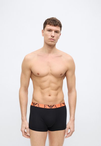 Άνδρας με κοντά, σγουρά μαλλιά που φοράει μαύρα ανδρικά εσώρουχα τύπου μποξερ με πορτοκαλί ζωνάρι μέσης και γράμματα λογότυπου, στέκεται μπροστά από απλό ανοιχτόχρωμο φόντο.