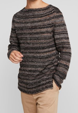 Mann trägt einen langärmeligen, gestrickten Pullover mit horizontalen schwarzen, grauen und braunen Streifen und beigen Hosen, steht mit einer Hand in der Tasche.