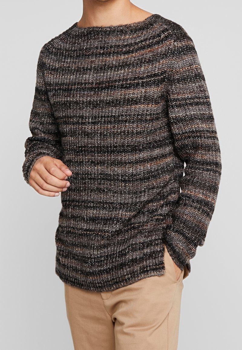 Homme portant un pull en maille à manches longues avec des rayures horizontales noires, grises et brunes et un pantalon beige, debout avec une main dans la poche.