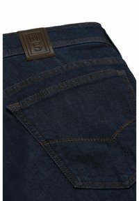 Jean denim bleu foncé avec un patch en cuir marron embossé "M5" et des coutures contrastantes orange le long des poches et des coutures.