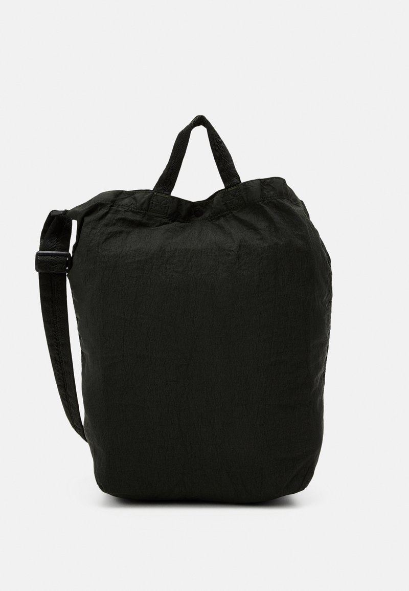 Sac cabas en tissu noir doux avec des poignées courtes et une longue bandoulière réglable sur un fond uni.
