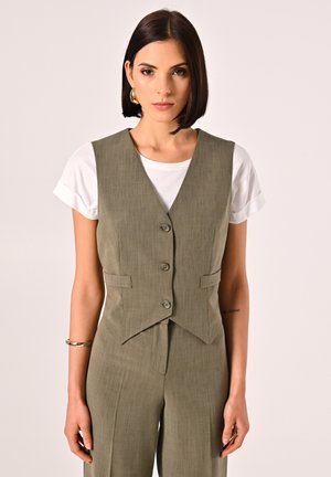 Gilet su misura verde oliva con colletto a scollo, tre bottoni e tasche frontali, indossato sopra una maglietta bianca; tessuto strutturato.