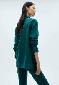 Teal oversized blouse met lange mouwen, knoopmanchetten en plooidetails aan de achterkant, gedragen over donkergroene slim-fit broek.
