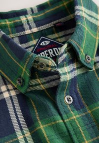 Zaļi un tumši zili rūtainas vilnas flaneļa kreklā ar apkakli, pogu aizdari un zīmola etiķeti. Audums ar tekstūru un kontrastējošām svītrām.