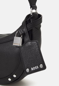 Černá texturovaná kožená kabelka se stříbrnými hroty, značkovým stříbrným zámkem a koženým štítkem s embosovaným nápisem "BOSS" v bílé barvě.