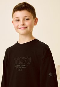 Svart bomullströja med "TOKYO" och koordinater tryckta i grått. Avslappnad passform med rund halsringning och långa ärmar.