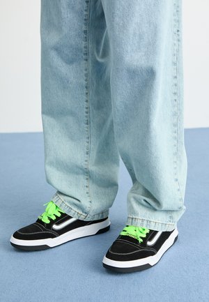Baskets noires avec des lacets vert fluo, tige en daim et mesh, semelle blanche et détails argentés. Portées avec un jean bleu clair ample.