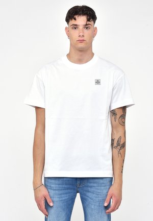 Hvid bomulds t-shirt med korte ærmer, rund hals og et lille rektangulært logopleth på venstre bryst. Båret med blå denim jeans.