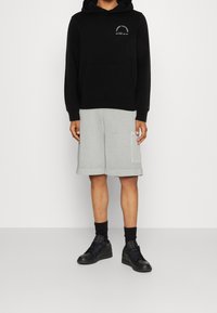 Sudadera negra con un logo en el pecho, combinada con pantalones cortos gris claro hasta la rodilla con bolsillos laterales y zapatillas deportivas negras.