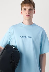 Lys blå bomulls t-skjorte med rund hals og korte ermer, prydet med et mørkeblått "Calvin Klein"-logo. Svart veske holdt i venstre arm.