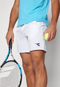 Valged tennisepüksid, millel on tumesinine logo, ning turgene särgid. Teises käes on tennisereket ja kollane pall.