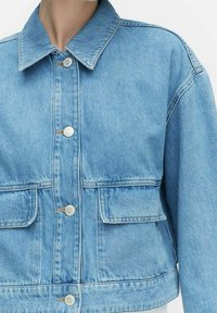 Vaaleansininen farkkutakki, jossa on kauluksellinen pääntie, kaksi etupussirakoa ja hopeiset napit. Lyhyt malli, jossa on hieman teksturoitu pinta.