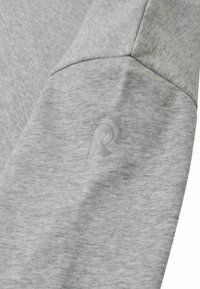 Szara rękawica z dresu z teksturowanej tkaniny, z delikatnym tłoczonym logo. Szwy podkreślają strukturę i design.