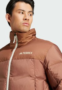 Brown insulated jacket with a high collar, zip closure, and textured fabric. Features a logo on the chest and contrasting lighter accents.