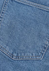 Tessuto di denim blu con una tasca posteriore appuntita e cuciture marroni a contrasto. La texture è leggermente ruvida con un aspetto sbiadito.