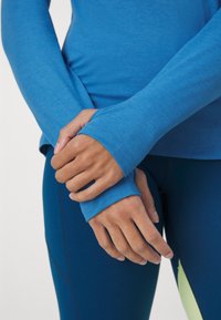 Modrá sportovní halenka s dlouhým rukávem a otvory pro palce, kombinovaná s modrými legínami. Materiál má hladkou strukturu a jemný melírovaný vzor.