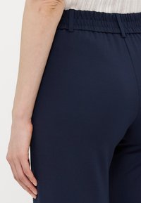 Navy blue pants with a stretchy waistband, featuring a smooth texture and a fitted design, shown from the side with a hand resting on the hip.