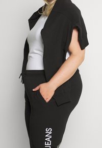 Schwarze cropped Jacke über einem weißen Tanktop, kombiniert mit schwarzen Jogginghosen mit einem weißen "JEANS"-Aufdruck. Beide aus einem weichen, bequemen Material gefertigt.