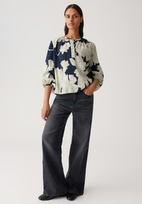 Vrouw in een losse bloeiende navy en witte blouse met 3/4 mouwen en een wijde, vervaagde zwarte jeans, staand met één hand in de zak.