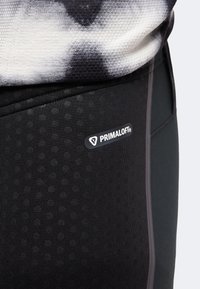 Gros plan sur des leggings en tissu noir avec le logo Primaloft sur la cuisse et une partie d'une chemise à motif noir et blanc au-dessus.