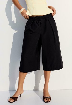 Osoba nosiaca čierne široké krátke nohavice, žltý tielkový top, biely náramok a hnedé sandále na podpätku, stojaca pri bielej stene.