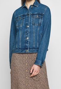 Denim jacket in medium blue with a classic cut, two chest pockets, silver hardware, and visible stitching details. Worn over a patterned skirt.
