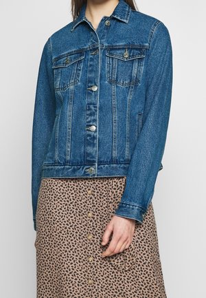 Veste en denim bleu moyen avec une coupe classique, deux poches poitrine, quincaillerie argentée et détails de couture visibles. Portée sur une jupe à motifs.