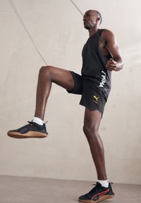 Zwarte sportieve tanktop en shorts met witte en gele accenten. Zwarte schoenen met een gumrubberen zool. Het onderwerp is aan het touwtjespringen, halverwege de beweging.